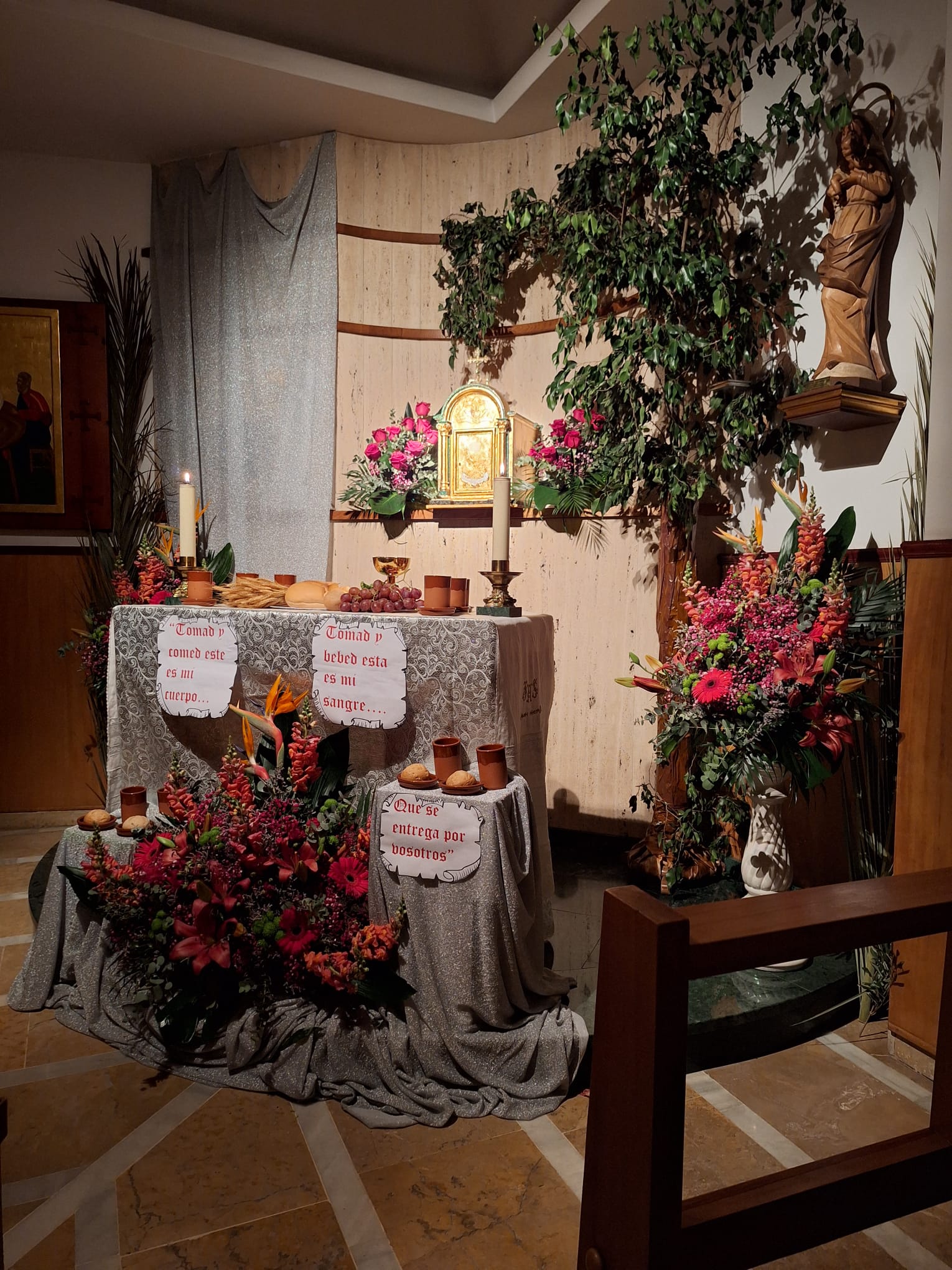 Imagen del sagrario en la capilla. El altar está decorado con panes y vasos de vino, uvas y trigo, y un cáliz, así como ramos de flores en el suelo y a los lados. También a los lados del sagrario hay dos ramos de rosas.