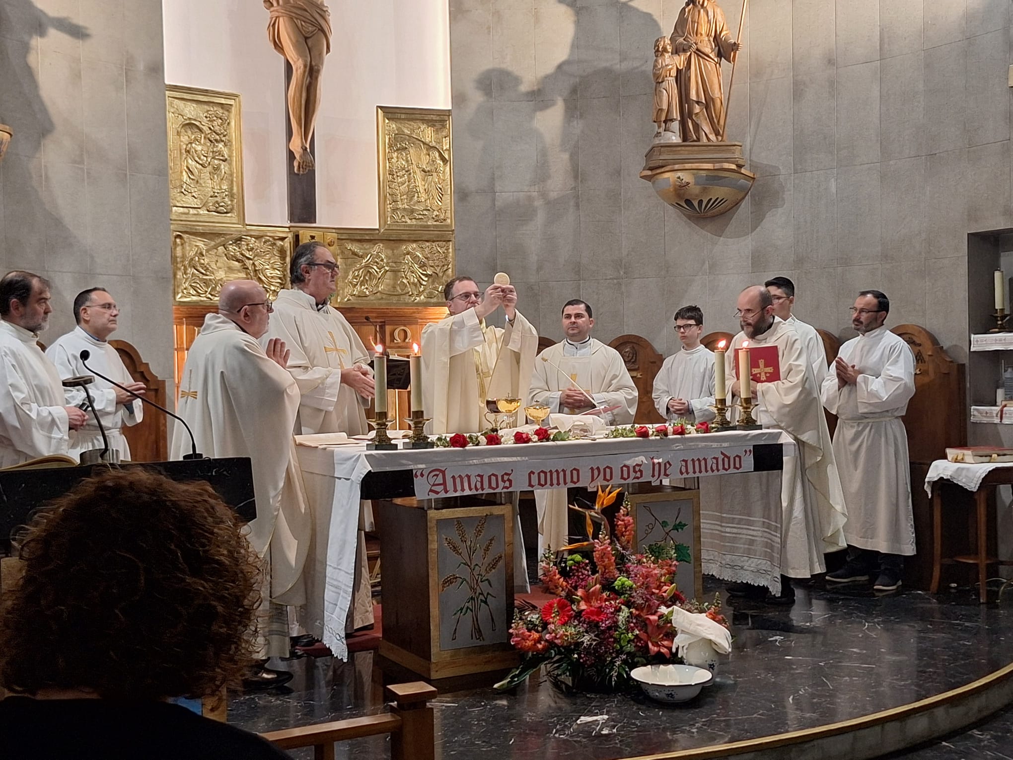 Fotografía tomada durante la consagración. José Manuel, el parróco, sostiene la forma consagrada, mostrándosela a los fieles. Le acompañan nuestro vicario parroquial, Juan Manuel, y los sacerdotes Greivin, Benjamín y Felipe. También en el altar están los acólitos. El altar está decorado con la frase "Amaos como yo os he amado", que se leyó en el evangelio del día. A sus pies, junto con un centro de flores, se encuentran la jofaina y la palangana con que José Manuel realizó el lavatorio de pies.